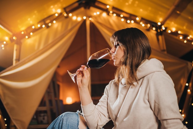 Close up of woman freelancer drinking wine sitting in cozy\
glamping tent in autumn evening luxury camping tent for outdoor\
holiday and vacation lifestyle concept