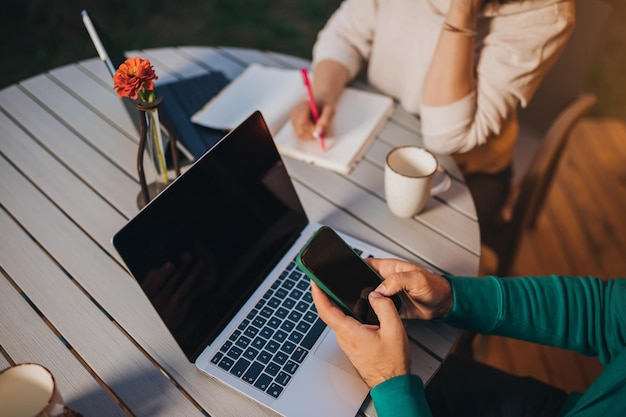 Close up of Woman freelancer working laptop and making notes while colleague using phone sitting in cozy glamping tent in summer evening