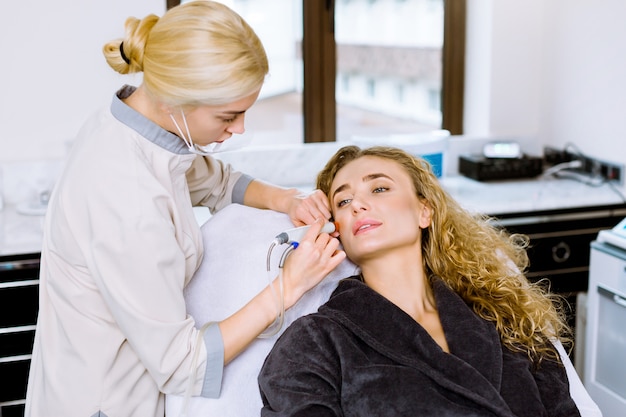 Close-up of woman getting facial hydro microdermabrasion peeling treatment and young female doctor beautician at beauty clinic. Hydra vacuum cleaner.