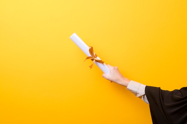 Close up of woman hand in graduation gown is holding graduation cap and certificate on yellow background