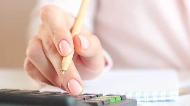 Close up of woman hand with calculator counting and taking notes to notebook. Finances, economy, technology concept. Pink background. Selective focus.