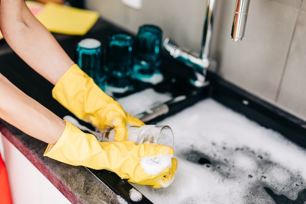 Primo piano della mano della donna in guanti di gomma protettivi gialli che lavano i piatti in cucina.