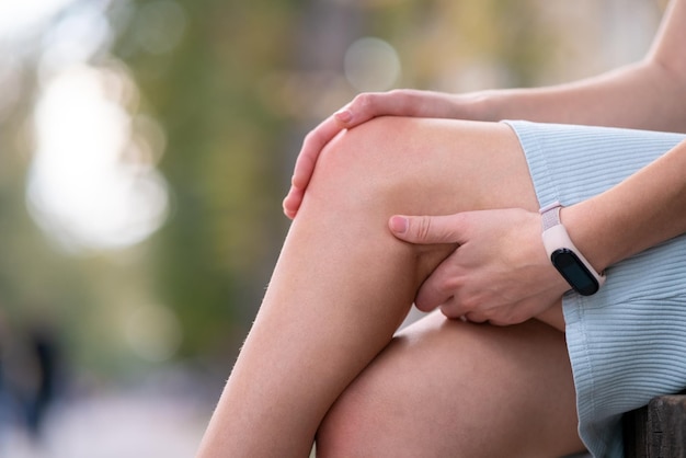 Primo piano delle mani della donna che tengono il ginocchio dolorante seduto sulla panchina all'aperto.