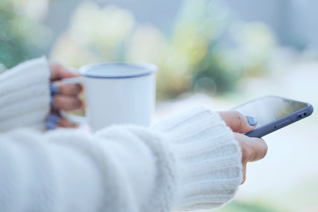 Close up woman hands holding hot cup of coffee or tea and using smartphone in cold weather background, wearing warm fur knitted clothes