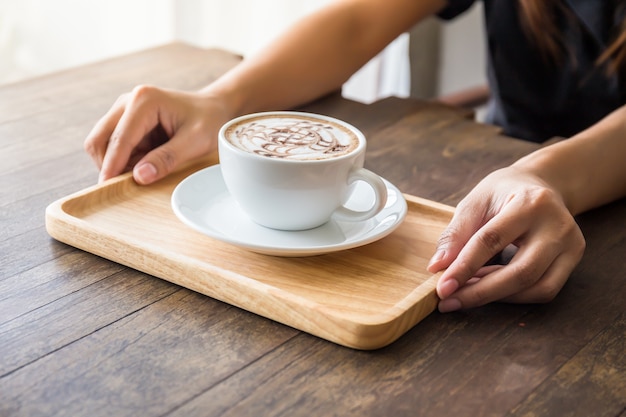 Close up of woman hands holding white cup of coffee with beautiful pattern art and wooden 