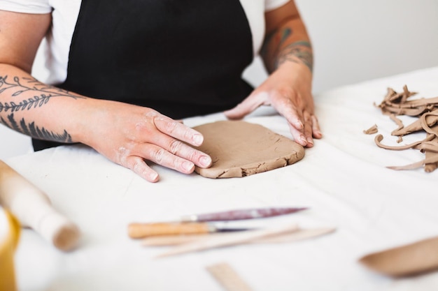 陶器のスタジオで粘土で作業している黒いエプロンの入れ墨で女性の手をクローズアップ
