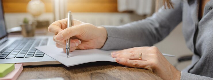 ノートパソコンの横にあるオフィスの机の上の紙に書くクローズアップの女性の手