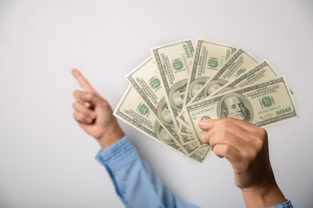 Close up woman holding dollars happy pointing with hand and finger to the side successful showing money and boasting lottery winning big profit indoor studio shot isolated on gray background