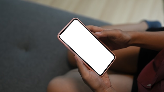 Close up of woman holding mobile phone with blank white screen while sitting on couch