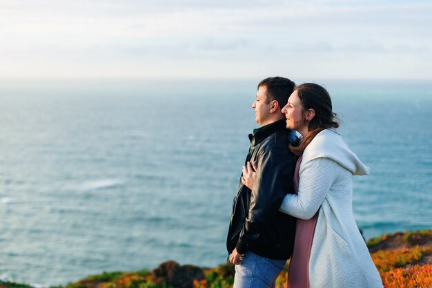 A close-up of a woman hugs her husband from the back side and they admire the ocean view