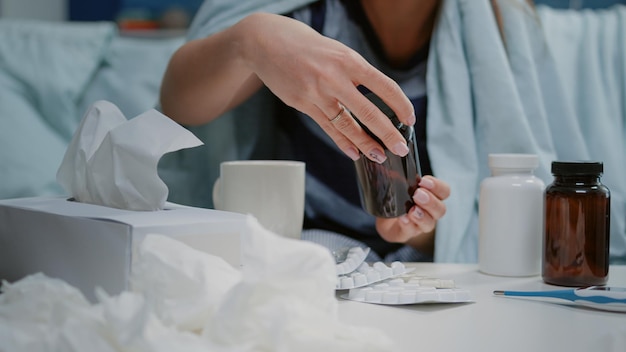 Close up of woman reading labels of tablets and jars with pills and capsules on table with medicaments, tissues and thermometer. Sick person looking for treatment to cure virus symptoms