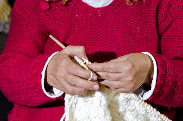 Close-up of woman in red sweater knitting white wool yarn with needles by hand.