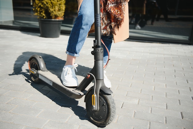 Chiuda in su della donna che guida il motorino di scossa elettrico nero al paesaggio urbano, sfocatura di movimento