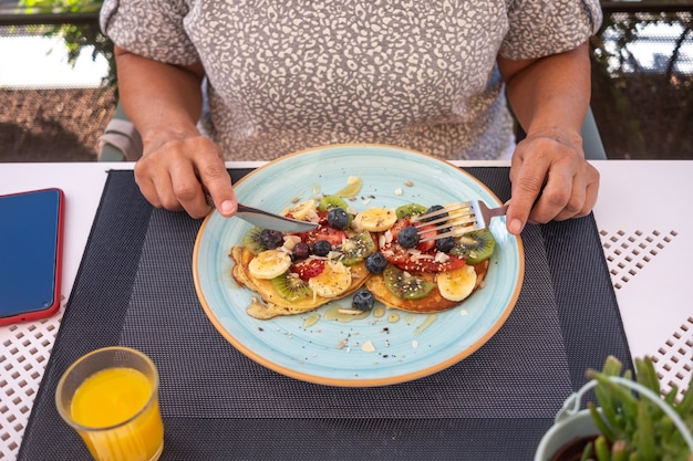 Close-up on woman\'s hands sitting at cafe table holding cutlery\
ready to eat an healthy breakfast, classic american pancakes with\
banana, kiwi, strawberry and honey. orange juice on the table