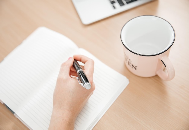 Close up of a woman's hands writing in a notepad placed on a wooden table cup for tea home office and work concept plans and thoughts