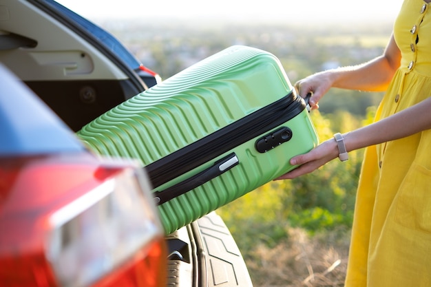 Close up of woman taking green suitcase from car trunk. Travel and vacations concept.