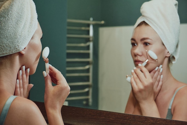 Close up woman using jade facial roller for face massage looking in the bathroom mirror