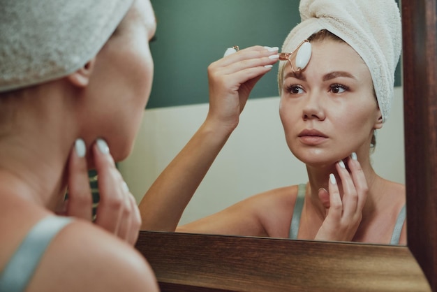 Крупным планом женщина, использующая нефритовый валик для лица для массажа лица, смотрит в зеркало в ванной