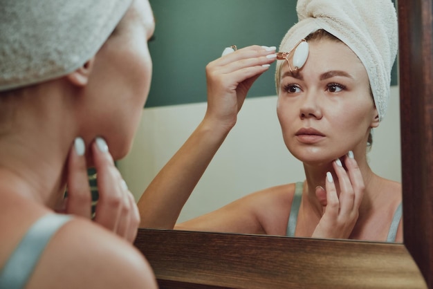 Close up woman using jade facial roller for face massage looking in the bathroom mirror