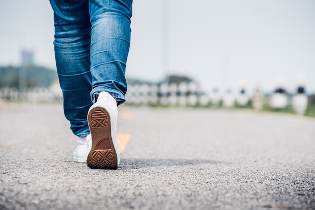 Chiuda sul jean di usura di donna e la scarpa da tennis bianca che cammina in avanti sulla strada della strada principale nel giorno soleggiato