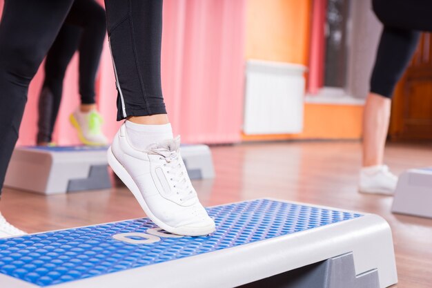 Close Up of Woman Wearing White Sneakers Doing Toe Tap on Step Platform in Aerobic Exercise Class with Group of Women in Background in Dance Studio