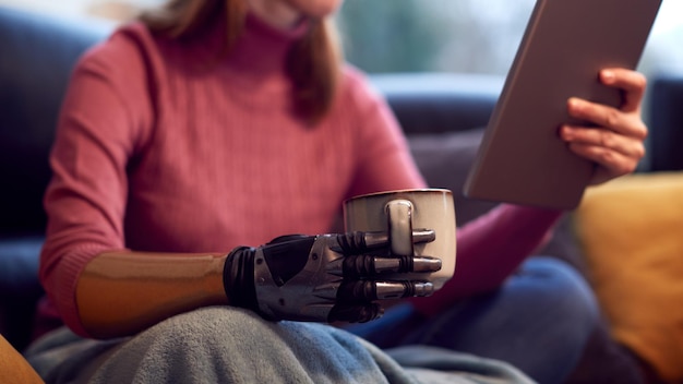 Foto primo piano di una donna con un braccio protesico e una mano a casa utilizzando una tavoletta digitale che beve una tazza di caffè