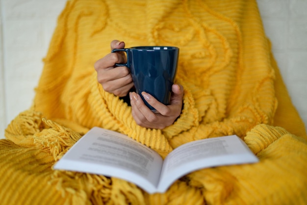 Close up of woman wrapped warm yellow blanket holding mug of coffee or tea and reading a book