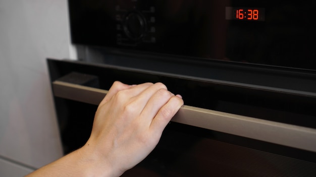 Foto primo piano della mano delle donne che apre la porta del forno per controllare l'arrosto. giovane donna che cucina in cucina aprendo la porta del forno