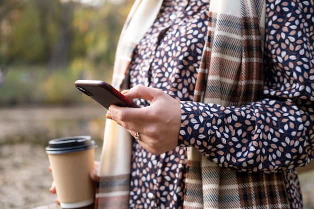 Close up women holding coffee cup using phone in nature in autumn park in fall