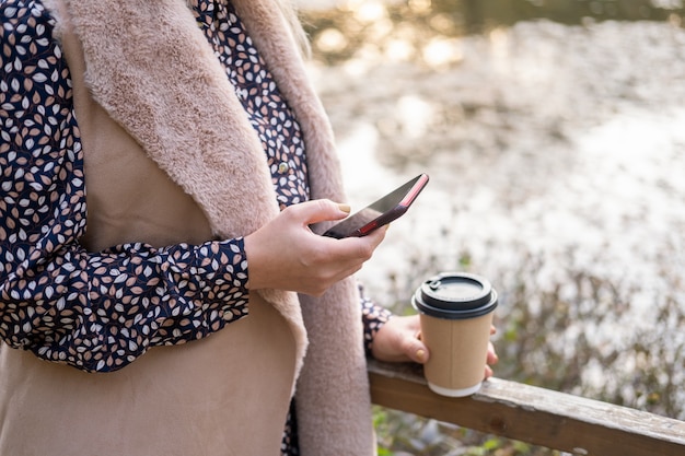 Close up women holding coffee cup using phone in nature in autumn park in fall