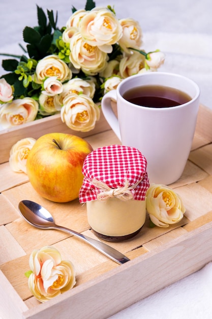 Close up of wooden tray with cup of tea, jam-jar, apple and flowers