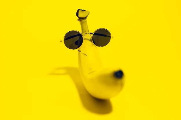 Close-up of yellow banana wearing round sunglasses lying on surface of yellow color.