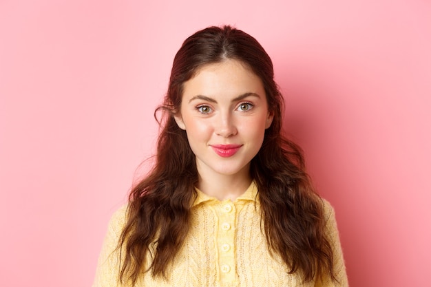 Close up of young 20s female model with cute feminine make up, smiling and looking hopeful at camera, standing against pink wall.