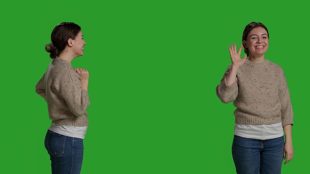 Photo close up of young adult greeting person with wave, smiling in studio. relaxed casual woman waving hi or hello on camera, standing over full body green screen background and saying goodbye.