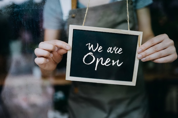close up of young asian female small business owner in apron starting her business day hanging an open sign on the door at the shop business owner and everyday business practice concept