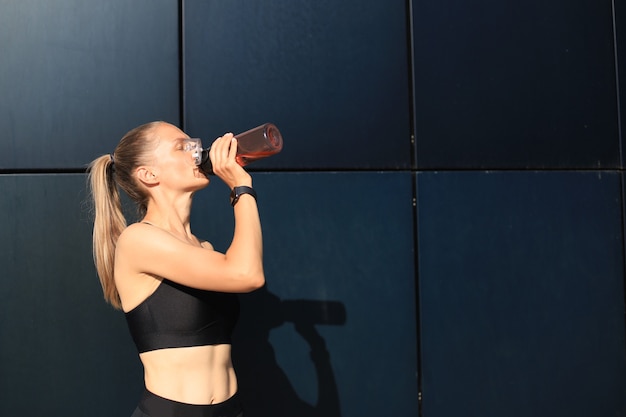 Close up of a young attractive tired sportswoman outdoors, drinking water from a bottle.