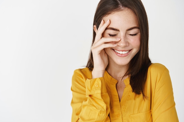 Primo piano di una giovane donna bruna che sorride felice, tocca la faccia perfetta senza trucco e ride con gli occhi chiusi, in piedi sul muro bianco