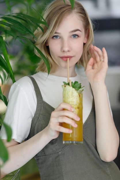 写真 居心地の良い緑のカフェで背景に大きな緑の葉とカメラで幸せそうに見てドレスを着た若い陽気な女性を閉じます。眼鏡の女の子。彼の手で飲み物を持って