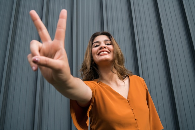 Close-up of young confident woman smiling and showing peace sign with fingers while standing outdoors on the street.