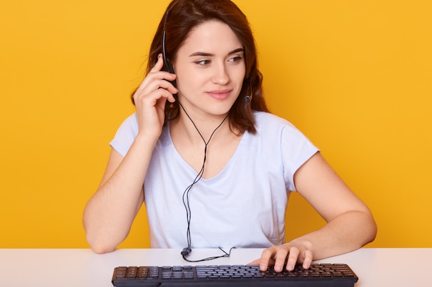 Фото Крупным планом молодая темноволосая девушка в наушниках с микрофоном, работает на клавиатуре и разговаривает с кем-то, имеет приятное и сосредоточенное выражение лица, носит белую футболку