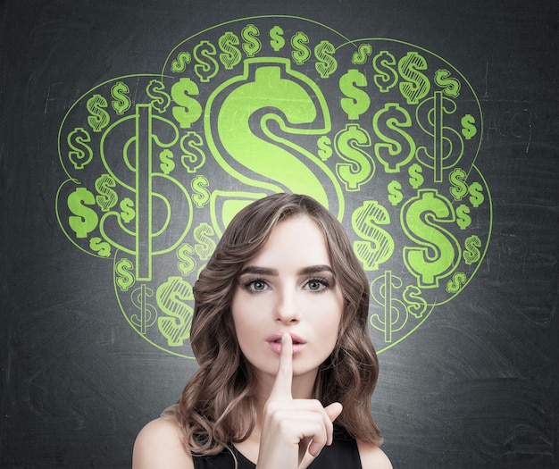 Close up of a young European woman with wavy hair making a hush sign. Blackboard background with a green dollar sign cloud sketch on it.