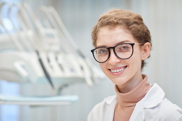 病院の正面に立って笑っている眼鏡の若い女性医師のクローズアップ