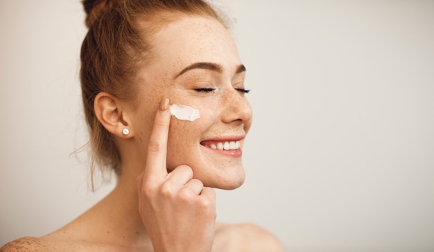 Photo close up of a young female with red hair and freckles applying white cream on her face laughing with closed eyes isolated on white wall.