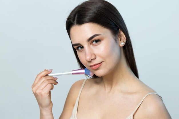 Close-up of a young girl in a light top on a white background making a facial make-up. A pretty woman holds a cosmetic brush near her face and smiles.