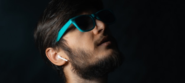 Close-up young hipster using wireless earphones, looking up wearing cyan sunglass, wall of black color.