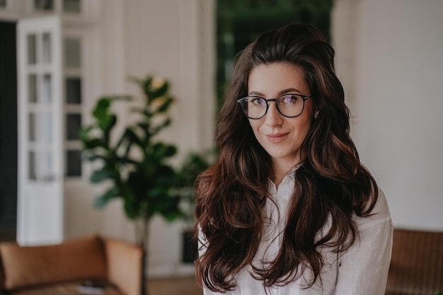 Close up young hispanic brunette woman in glasses with wavy loose hair in white shirt looking at camera adorable smiling over blurry office Portrait of smart student at home Business and education