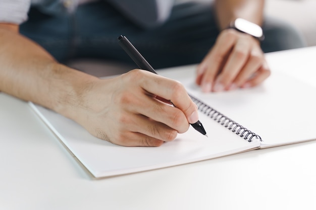 Close up of young man in casual cloth hands writing down on the notepad, notebook using ballpoint pen on the table.