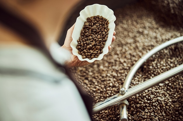 Foto primo piano di un giovane che controlla la qualità dei chicchi di caffè mentre l'uomo in piedi vicino alla macchina per la torrefazione del caffè