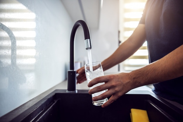 Close up of young man pouring fresh water from kitchen sink. Home interior.