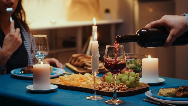 Photo close up of young man pouring red wine in wife glass. romantic caucasian happy couple sitting at the table in kitchen celebrating with candle lights, love and anniversary. romance surprise in relation